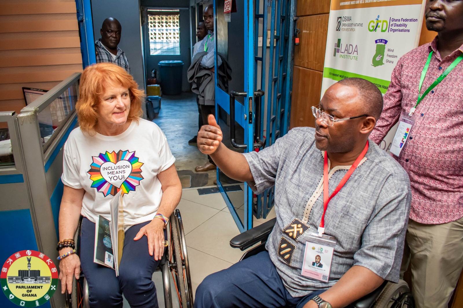 A woman wearing a white T-shirt with the slogan 'Inclusion Means You' speaks to a man wearing glasses and a grey shirt. Both are wheelchair users.